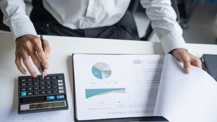 Beatrix Konnect A person in a white shirt uses a calculator with one hand and flips through financial reports with graphs and charts on a desk with the other hand.